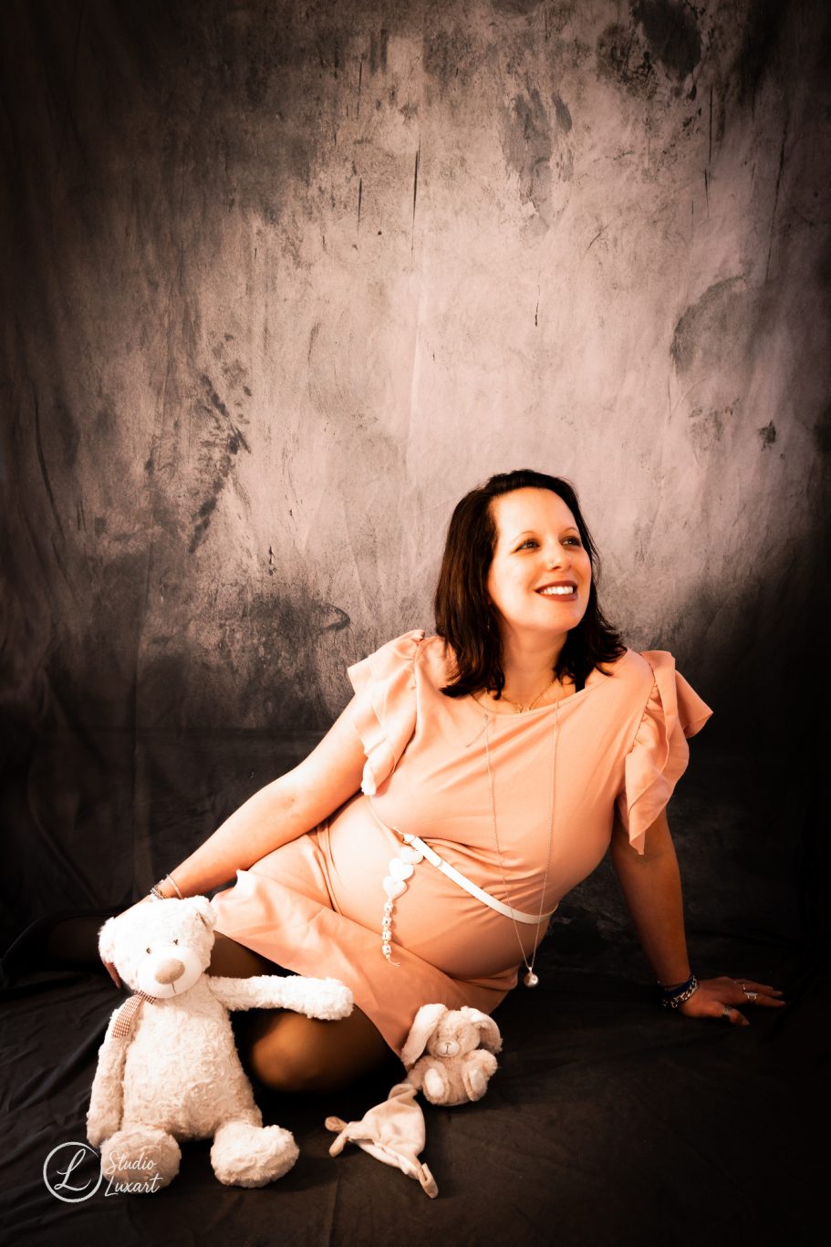 Femme souriante en robe rose, entourée de peluches blanches sur fond sombre.