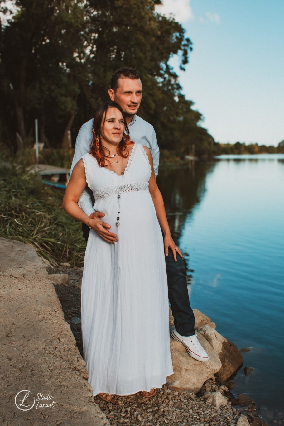Un couple souriant près d'un lac, la femme enceinte en robe blanche.